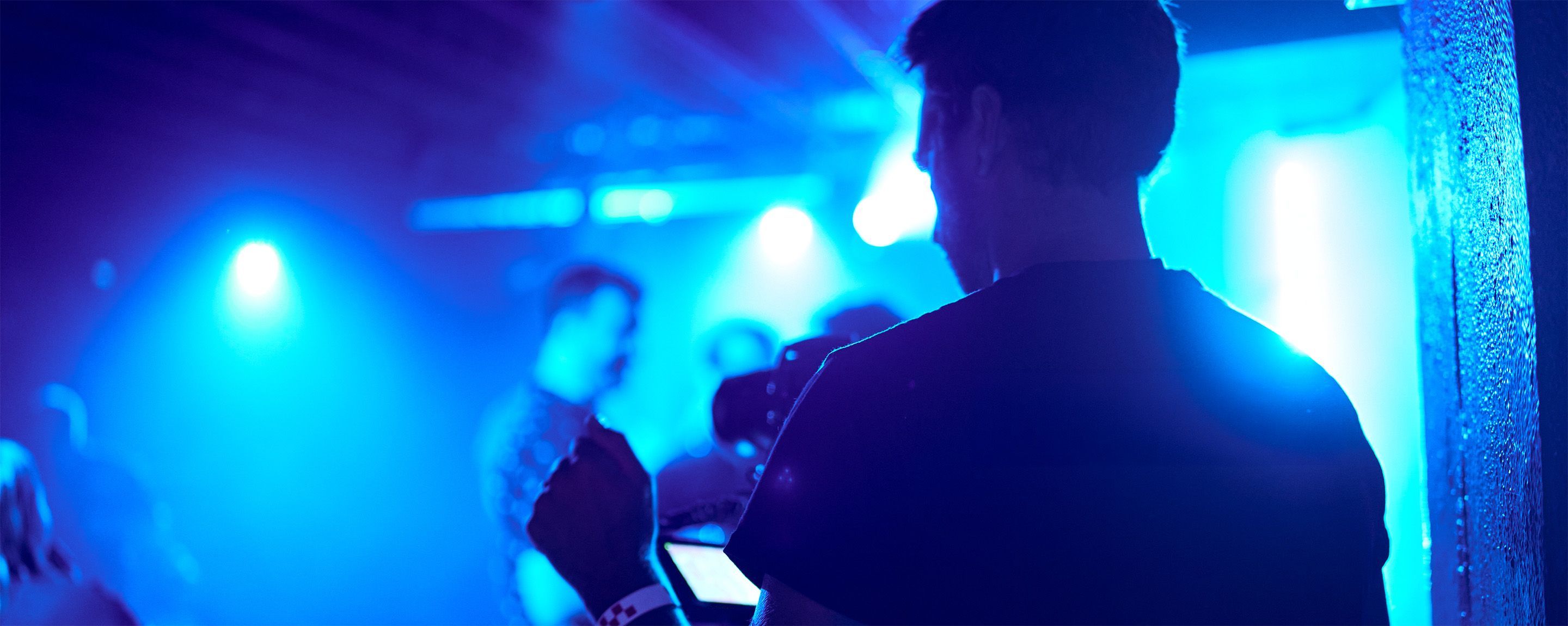 Silhouetted person holding a camera or video equipment in a dimly lit room with blue stage lights and blurry figures in the background.